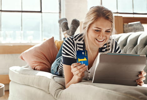 Smiling woman reclined on couch with holding a smart tablet in one hand an a Golden 1 credit card in the other