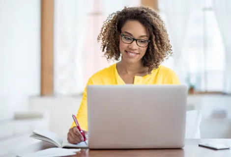 Woman looking at her laptop and writing on a notepad.