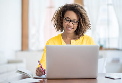 Woman looking at her laptop and writing on a notepad.