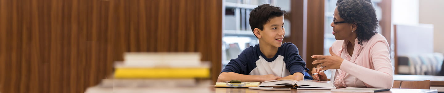 A woman and young boy sitting at a library table with a book in front of them engaged in discussion