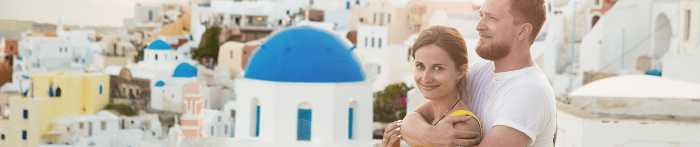Smiling man standing behind smiling woman with his arms around her with a Greek town in the background
