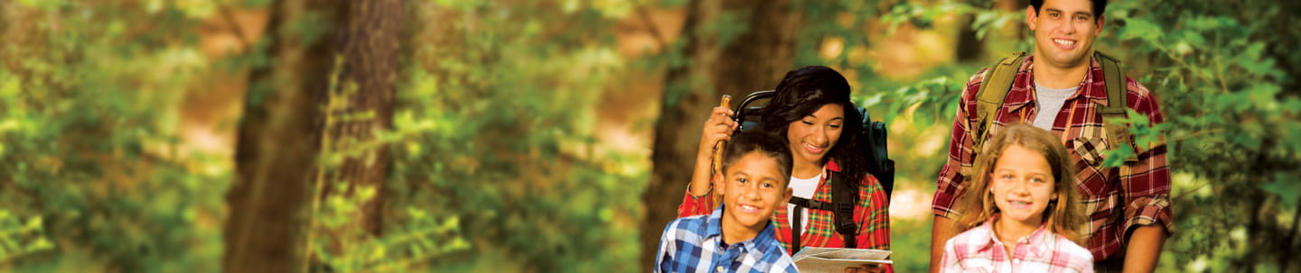 Smiling man and woman wearing backpacks, and boy and girl all hiking in woods; woman is holding walking stick and a map