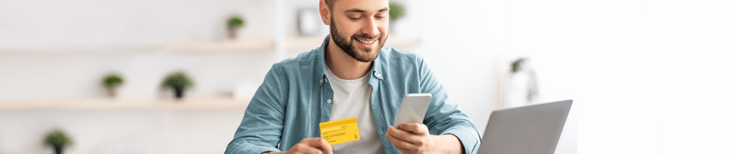 Smiling man sitting in front of laptop holding card in one hand and smartphone in the other