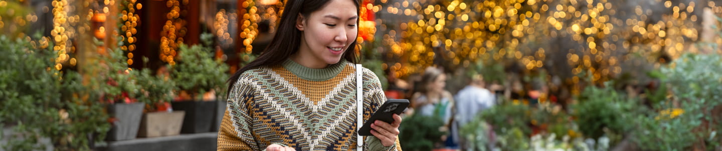 Woman in sweater looking at phone in front of store with Christmas lights