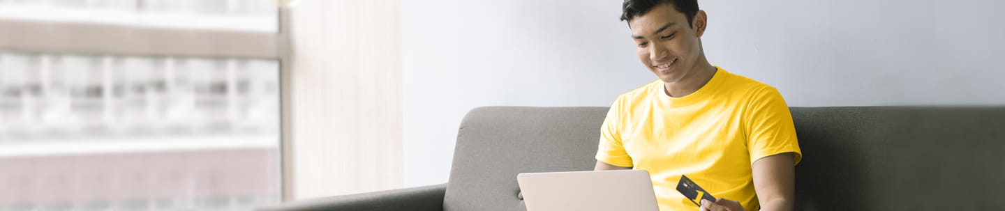 Smiling young man sitting on couch with Golden 1 credit card in one hand looking down at tablet device in his lap