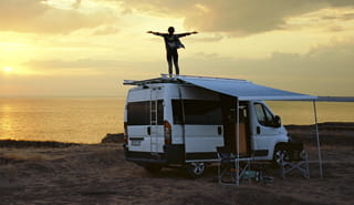 Person standing on roof of RV arms outstretched facing the sun setting over a body of water