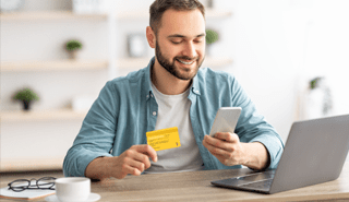 Smiling man sitting in front of laptop holding card in one hand and smartphone in the other