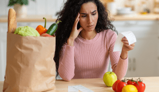 Woman holding her head, with a full grocery bag next to her, frowning at a grocery receipt